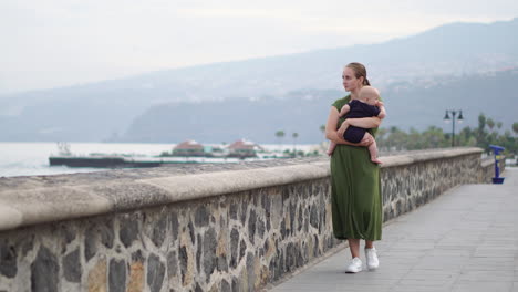 Walking-near-the-ocean-on-an-old-European-square,-a-young-mother-enjoys-the-presence-of-her-baby-son,-looking-at-the-waves-and-smiling-with-affection